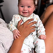 Margot is registered to the contest to win money with this photo: baby, child, smiling, person, hand, tattoo, pajamas, floral_pattern, indoor, couch, seated, happy, face, skin, arm, legs, cozy, blurred_background, infant, holding