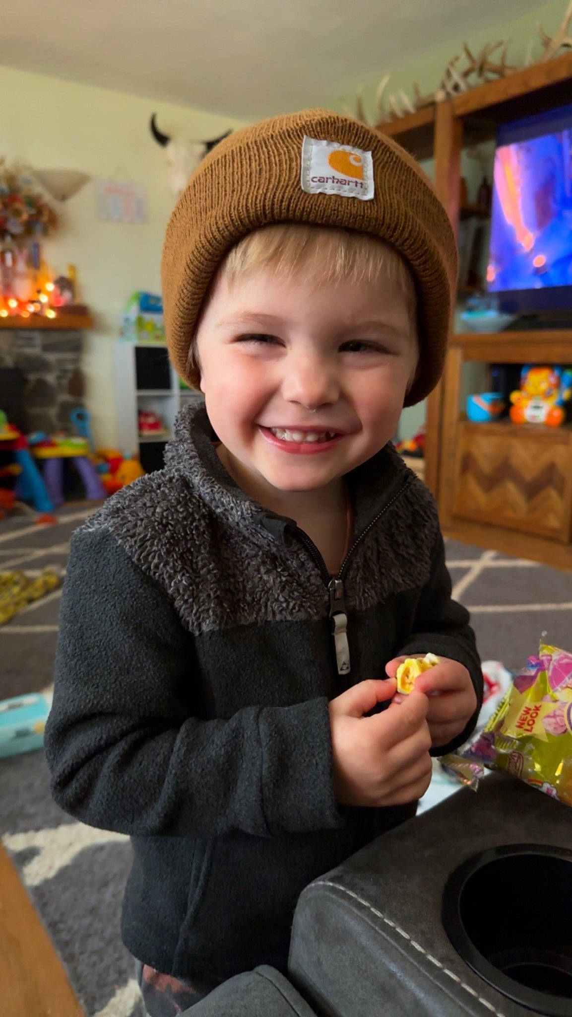 Kai is registered to the contest to win money with this photo: beanie, cap, child, event, facial_expression, fashion_accessory, fun, happy, headwear, holiday, jacket, joy, knit_cap, person, sitting, skin, smile, television, toddler, tree