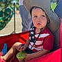 toddler, child, hat, stroller, red, outdoor, sunlight, blue_eyes, snack, container, shirt, stars, stripes, person, summer, casual, curious, seated, daytime, portrait