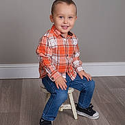 Andrew is registered to the contest to win money with this photo: child, toddler, smiling, plaid_shirt, orange_shirt, blue_jeans, sneakers, black_shoes, sitting, stool, wooden_floor, indoor, portrait, cute, happy, boy, short_hair, casual_clothing, studio_photo, neutral_background