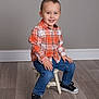 child, toddler, smiling, plaid_shirt, orange_shirt, blue_jeans, sneakers, black_shoes, sitting, stool, wooden_floor, indoor, portrait, cute, happy, boy, short_hair, casual_clothing, studio_photo, neutral_background