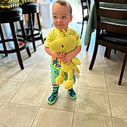 Matthew is registered to the contest to win money with this photo: toddler, child, plush_toy, yellow, indoor, floor, tile, clothing, rain_boots, curious, expression, furniture, chair, stool, kitchen, person, toy, standing, looking_sideways, playful
