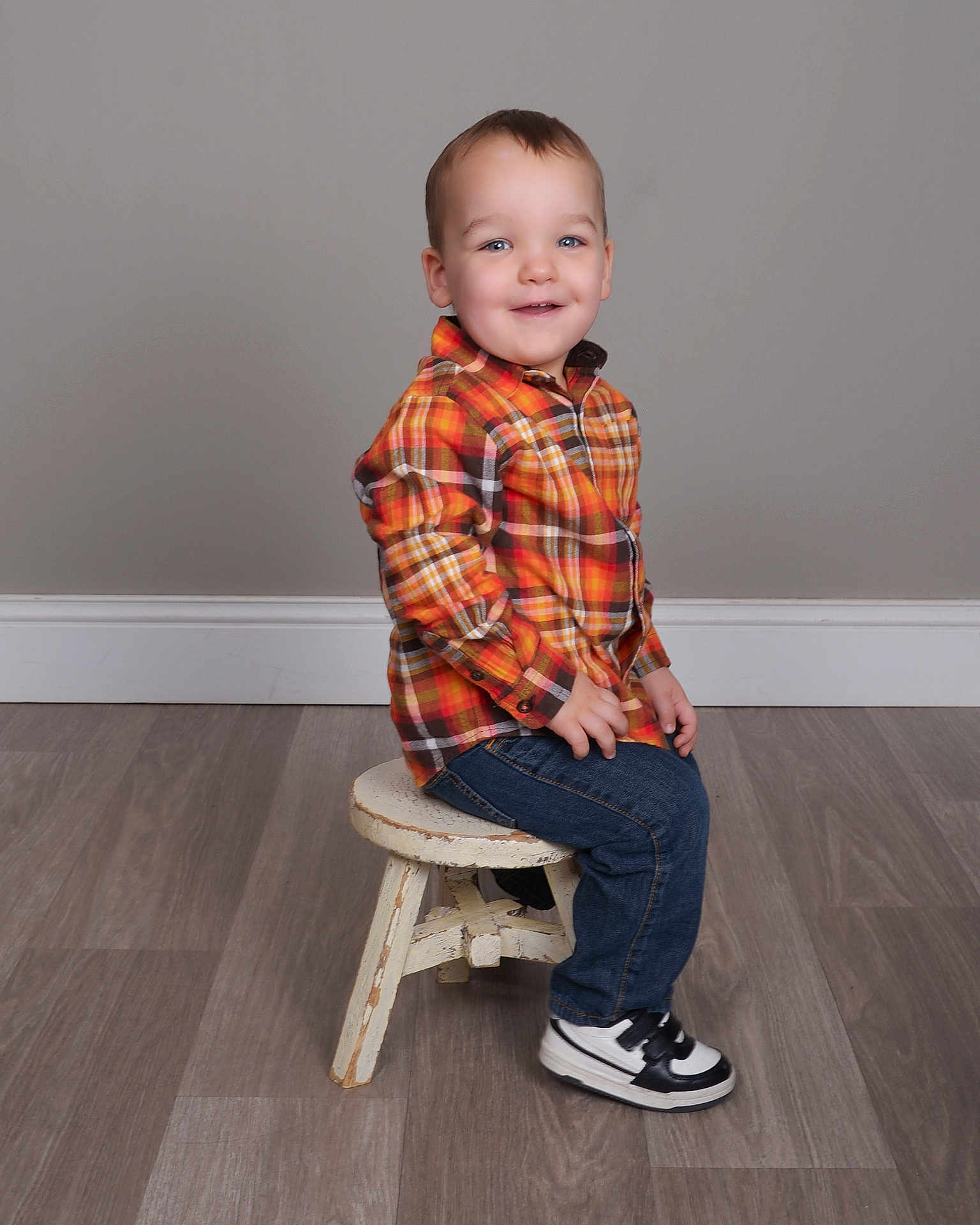 Matthew is registered to the contest to win money with this photo: toddler, child, smiling, plaid_shirt, jeans, sneakers, stool, indoor, portrait, happy, casual_clothing, floor, wooden_floor, gray_wall, seated, young_child, cute, studio, small_person, boy