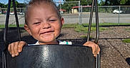 Cain is registered to the contest to win money with this photo: child, toddler, swing, playground, smiling, happy, outdoor, sunny, blue_sky, clouds, metal_chains, fence, trees, grass, clothing, face, hands, seat, daytime, fun