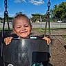 child, toddler, swing, playground, smiling, happy, outdoor, sunny, blue_sky, clouds, metal_chains, fence, trees, grass, clothing, face, hands, seat, daytime, fun