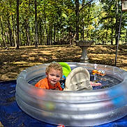 Cain is registered to the contest to win money with this photo: backyard, birdbath, blue_tarp, child, curly_hair, forest, inflatable_pool, orange_shirt, outdoor, pool, pool_toys, portrait, summer, sunlight, swimming, toddler, toy_boat, toy_car, trees, water