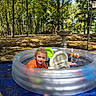 backyard, birdbath, blue_tarp, child, curly_hair, forest, inflatable_pool, orange_shirt, outdoor, pool, pool_toys, portrait, summer, sunlight, swimming, toddler, toy_boat, toy_car, trees, water