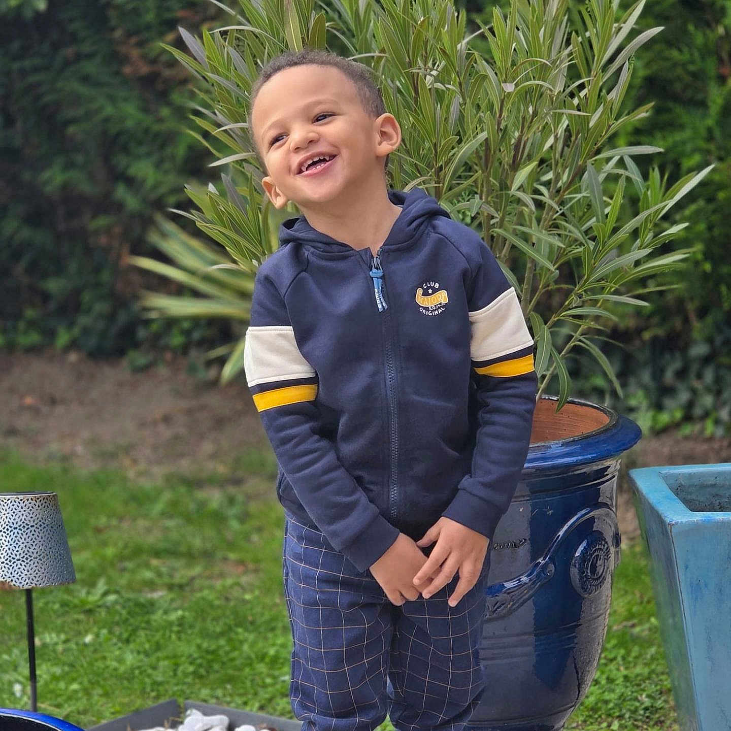Mahé a rejoint le concours — aidez-le/la à gagner de superbes lots ! boy, casual_clothing, child, cute, daylight, garden, grass, greenery, happy, hoodie, joyful, laughter, nature, outdoor, pants, plant, portrait, pot, smiling, standing