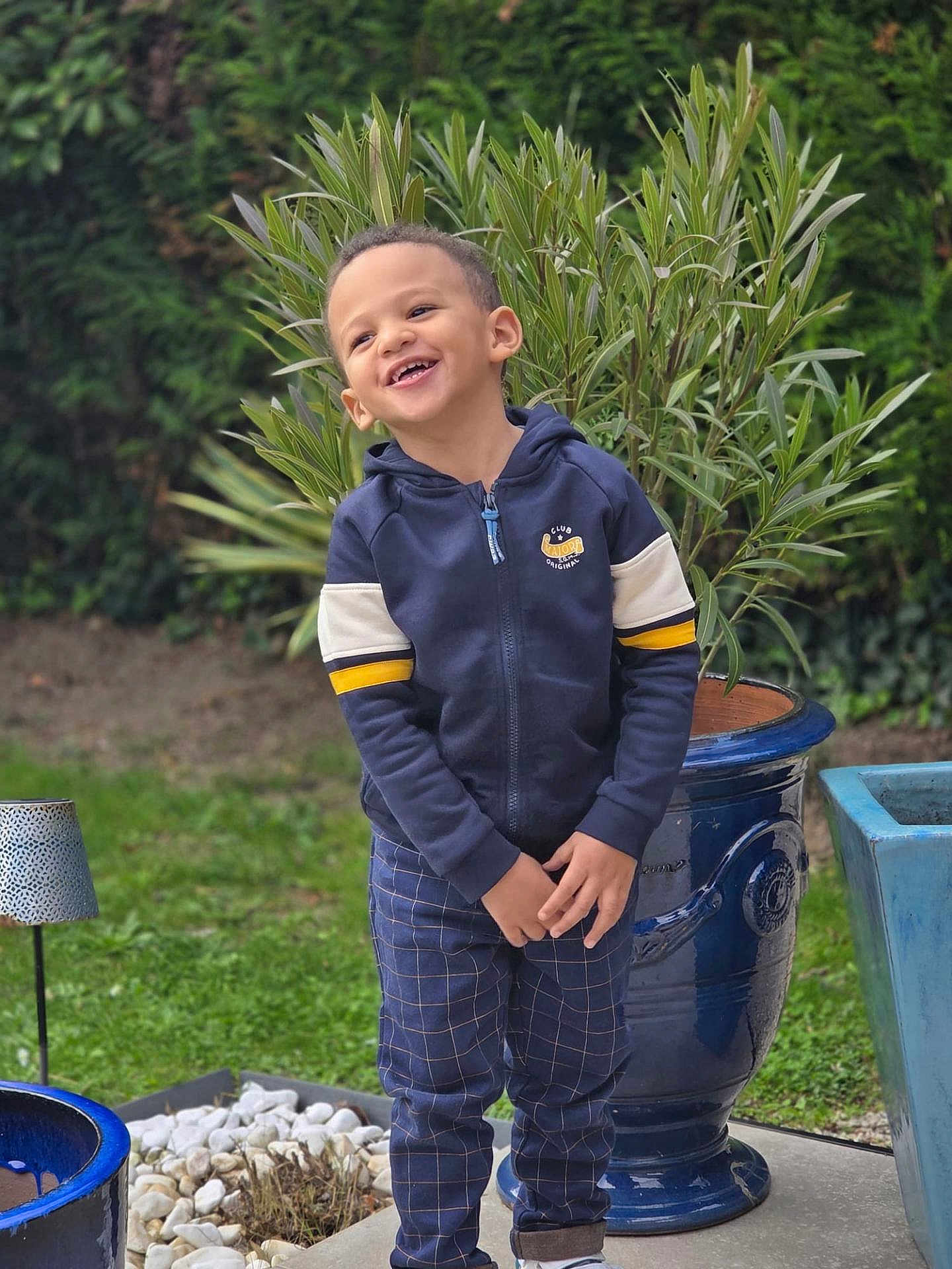Mahé a rejoint le concours — aidez-le/la à gagner de superbes lots ! child, boy, smiling, outdoor, plant, greenery, pot, garden, happy, casual_clothing, hoodie, pants, standing, nature, grass, daylight, portrait, cute, joyful, laughter
