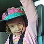child, hat, car_seat, pink_clothing, smiling, seatbelt, floral_pattern, raising_hand, indoors, young_child, face, person, casual, playful, happy, seat, straps, colorful, portrait, kid