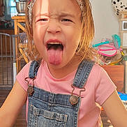 Taytum joined the competition — help win amazing prizes! child, tongue_out, denim_overalls, pink_shirt, headband, flower, ribbons, indoor, wooden_floor, playful, mischievous_expression, person, young_child, sunlight, face, hair, clothing, home, cute, happy