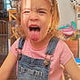 child, tongue_out, denim_overalls, pink_shirt, headband, flower, ribbons, indoor, wooden_floor, playful, mischievous_expression, person, young_child, sunlight, face, hair, clothing, home, cute, happy