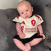 Luca is registered to the contest to win money with this photo: baby, infant, child, person, sitting, couch, sofa, clothing, jersey, football, sportswear, red_eye, indoor, soft_lighting, head_tilt, cute, feet, skin, portrait, home