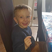 Nolan participe au concours pour gagner de l'argent avec cette photo : child, smiling, indoor, chair, table, tablecloth, person, sweater, pants, happy, gesture, face, eyes, hair, furniture, blurred_background, light, wooden_floor, casual_clothing, home