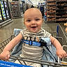 baby, smiling, shopping_cart, grocery_store, child, striped_clothing, blue_blanket, seatbelt, aisle, shelves, products, floor, person, head, hand, happy, indoor, retail, infant, cute
