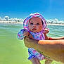 Abby is registered to the contest to win money with this photo: baby, child, water, beach, sunhat, swimsuit, dinosaur_pattern, tattoo, arms, holding, sky, clouds, water_reflection, sunlight, summer, outdoor, nature, person, portrait, vacation