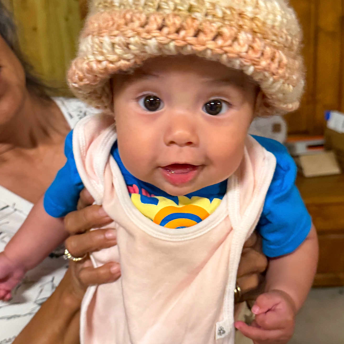Nayeli joined the competition — help win amazing prizes! baby, child, knit_hat, blue_shirt, bib, hands, person, wooden_background, indoor, cute, face, eyes, smiling, infant, clothing, portrait, warm_lighting, holding, expression, cozy