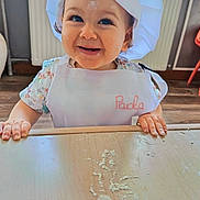 Paola a rejoint le concours — aidez-le/la à gagner de superbes lots ! baby, child, apron, chef_hat, flour, table, smiling, face, hands, indoor, kitchen, cooking, cute, person, young_child, wood_floor, radiator, white_clothing, playful, portrait