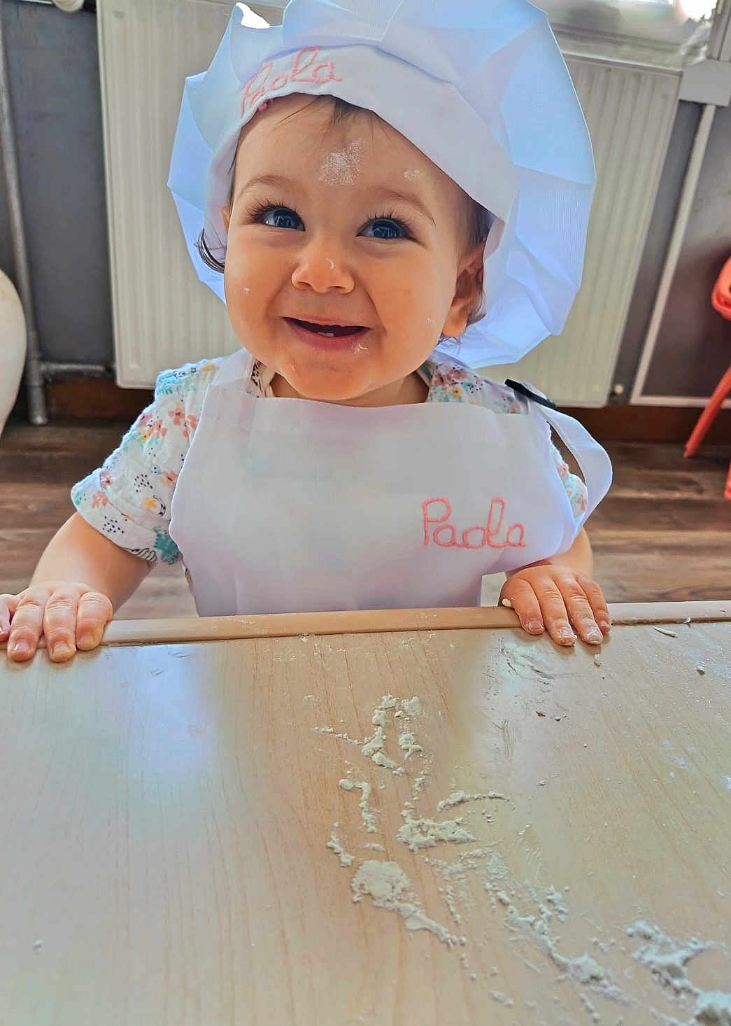 Paola a rejoint le concours — aidez-le/la à gagner de superbes lots ! baby, child, apron, chef_hat, flour, table, smiling, face, hands, indoor, kitchen, cooking, cute, person, young_child, wood_floor, radiator, white_clothing, playful, portrait