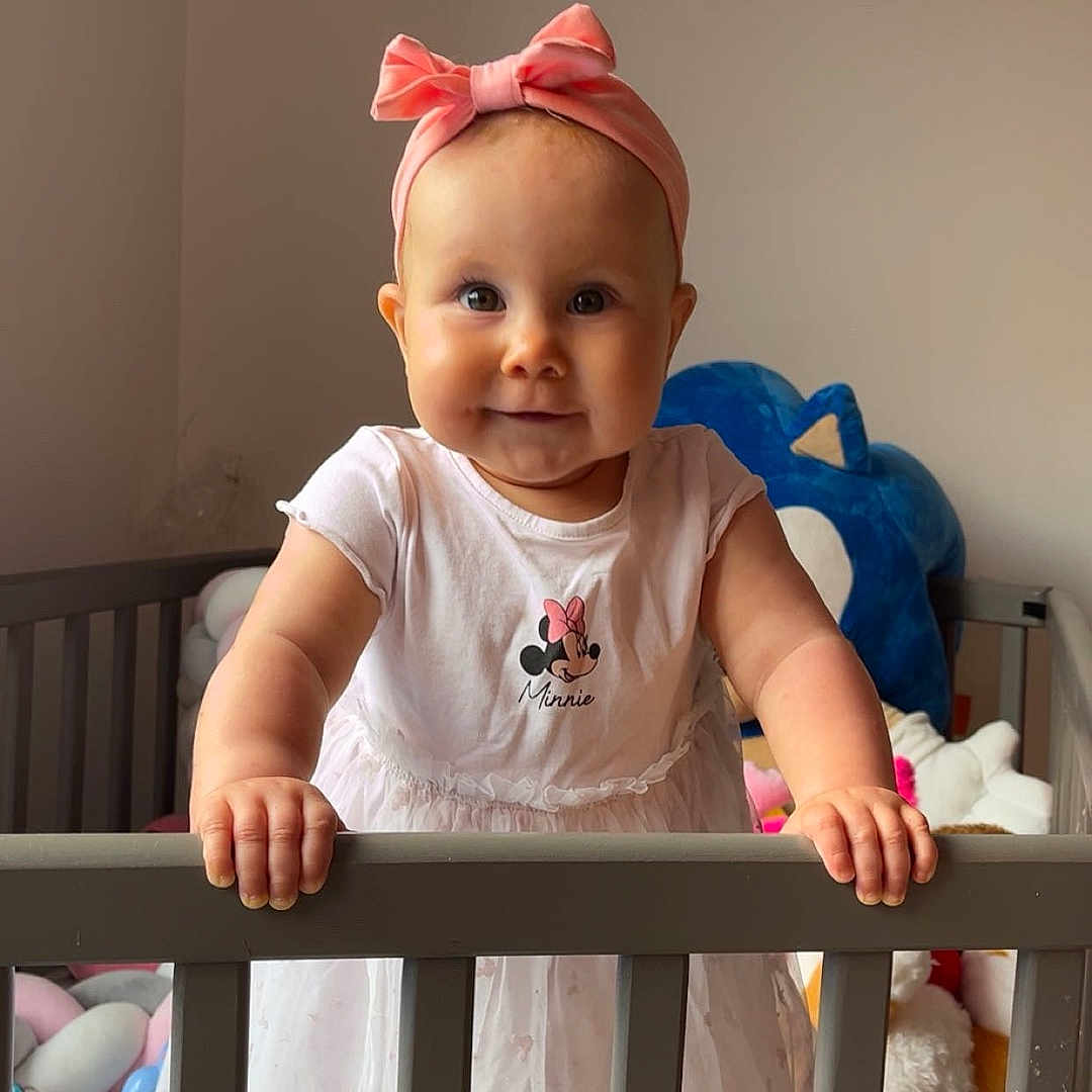 Julliana participe au concours pour gagner de l'argent avec cette photo : baby, child, crib, cute, dress, face, hand, happy, headband, indoor, infant, mickey_mouse, person, pink_bow, plush_toys, smile, standing, stuffed_animals, toy, white_dress