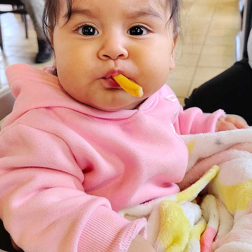Lia joined the competition — help win amazing prizes! baby, blanket, child, cozy, cute, expression, face, food, french_fry, hair, hand, high_chair, indoor, person, pink_hoodie, restaurant, seated, snack, tile_floor, young_child