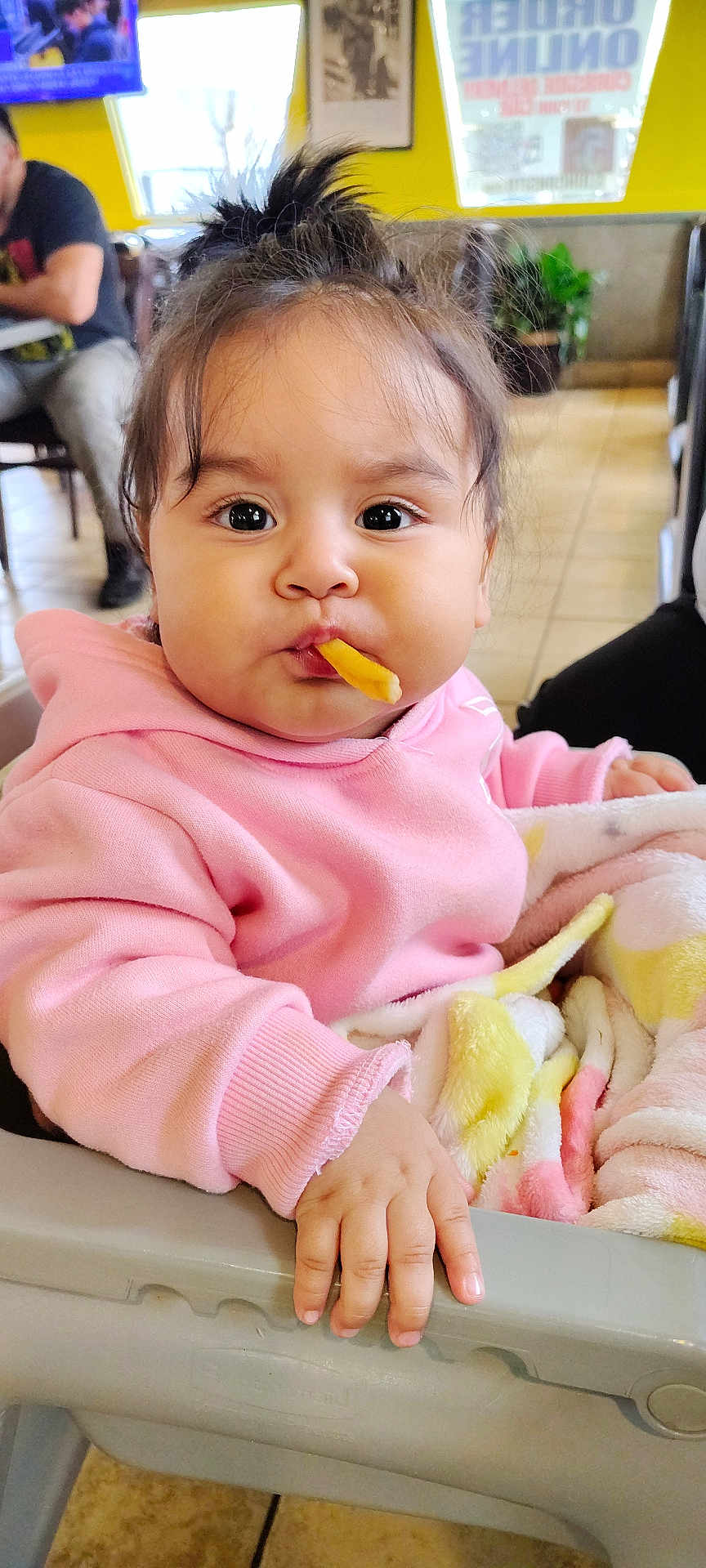 Lia joined the competition — help win amazing prizes! baby, child, face, french_fry, food, pink_hoodie, blanket, high_chair, hand, indoor, restaurant, tile_floor, person, cute, young_child, expression, snack, seated, hair, cozy