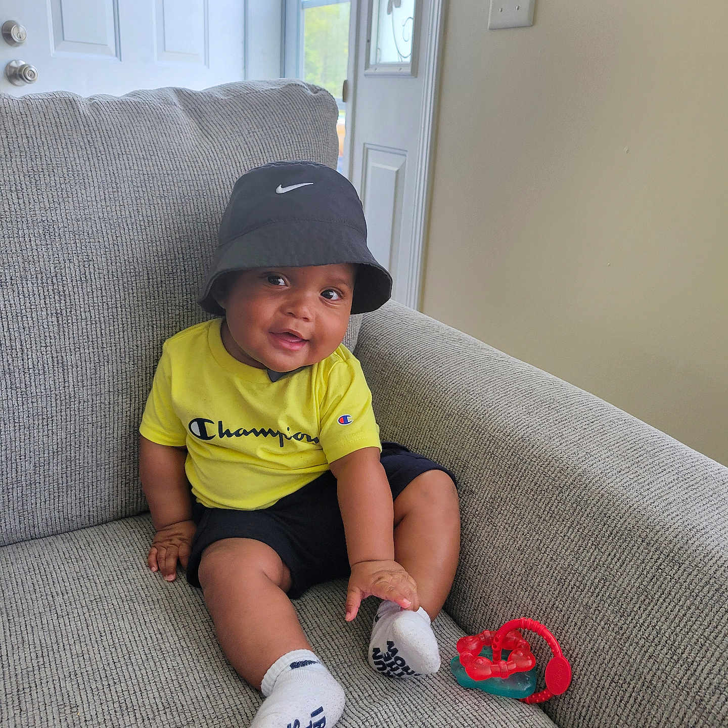 Darien joined the competition — help win amazing prizes! baby, bucket_hat, casual_clothing, champion, child, couch, door, furniture, happy, hat, home_interior, indoor, infant, light_switch, person, smiling, socks, teething_toy, window, yellow_shirt