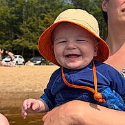 Vincent joined the competition — help win amazing prizes! baby, child, smiling, orange_hat, blue_clothing, adult_hand, beach, outdoor, sunny, water, trees, sand, person, portrait, happy, summer, daylight, nature, holding, casual