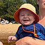 baby, child, smiling, orange_hat, blue_clothing, adult_hand, beach, outdoor, sunny, water, trees, sand, person, portrait, happy, summer, daylight, nature, holding, casual