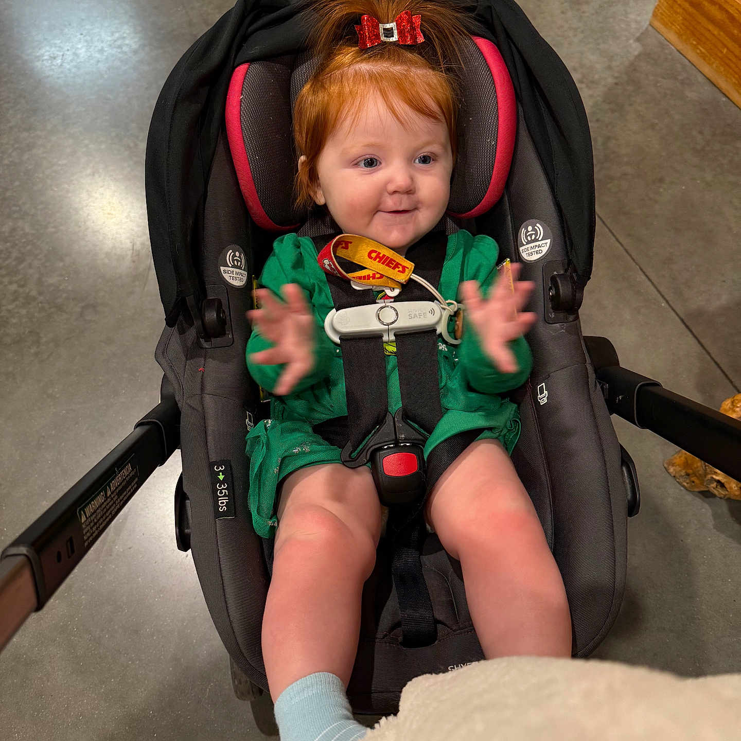 Evalyna is registered to the contest to win money with this photo: baby, car_seat, child, clapping, concrete, cute, expression, floor, footwear, green_dress, happy, indoor, infant, legs, person, red_bow, seatbelt, smile, strap, toddler