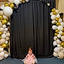 baby, child, pink_dress, balloons, white_balloons, gold_balloons, black_feathers, roses, black_curtain, blue_carpet, indoor, celebration, smiling, sitting, decor, party, cute, young_child, event, portrait