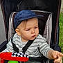 baby, cap, child, clothing, colorful, expression, grass, hand, headwear, outdoor, pavement, person, play, pointing, seatbelt, stroller, toddler, toy, transport, wheel