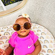 Cynthia is registered to the contest to win money with this photo: baby, child, sunglasses, headband, pink_shirt, floral_skirt, rainbow_pattern, cushion, indoor, plant, decor, fashion, cute, portrait, infant, smiling, accessory, colorful, happy, cozy