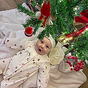 Dohya a rejoint le concours — aidez-le/la à gagner de superbes lots ! baby, infant, christmas_tree, ornaments, holiday_decor, blanket, white_clothing, headband, floor, wood_floor, greenery, bells, red_ornaments, lights, festive, cute, indoors, celebration, person, holiday