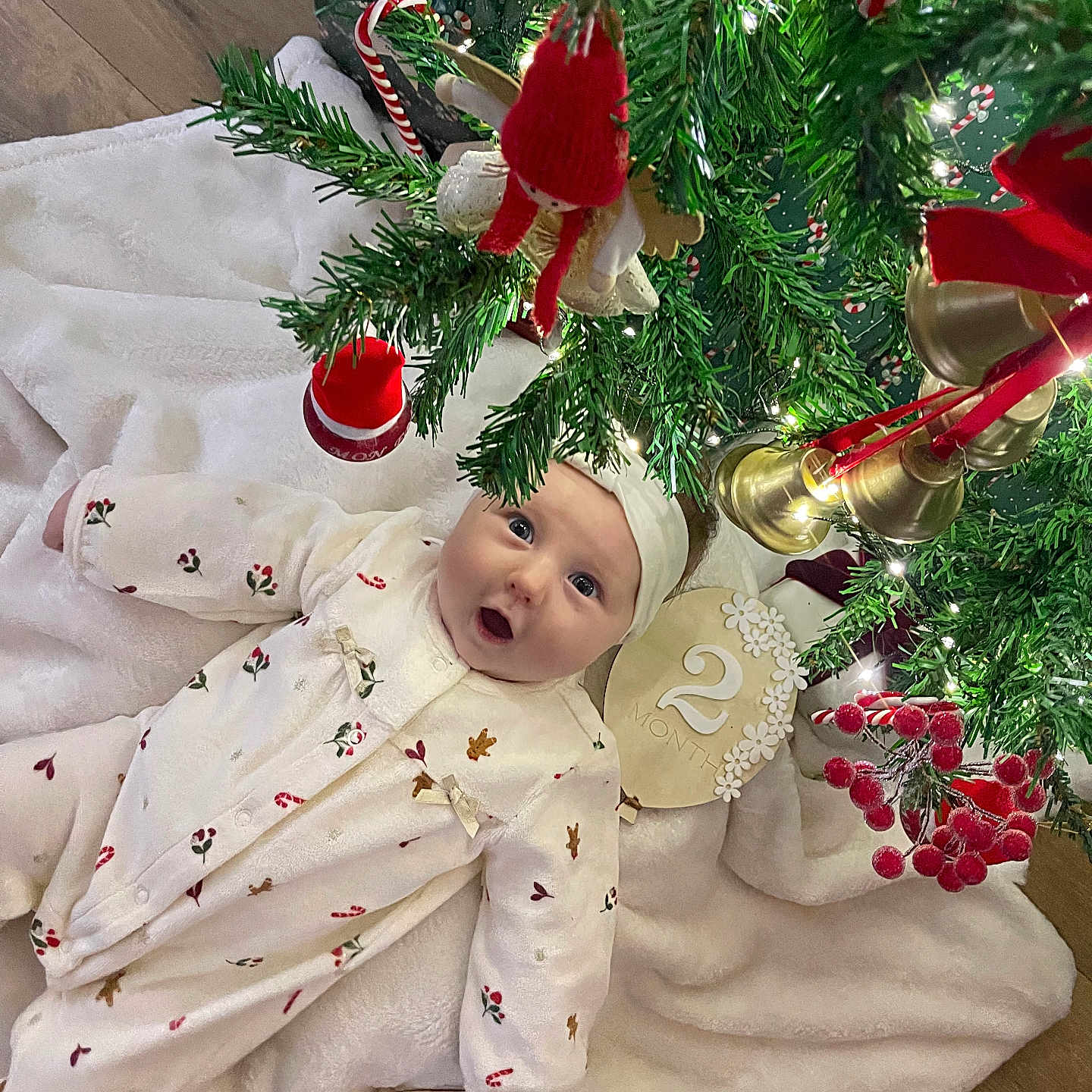 Dohya a rejoint le concours — aidez-le/la à gagner de superbes lots ! baby, bells, blanket, celebration, christmas_tree, cute, festive, floor, greenery, headband, holiday, holiday_decor, indoors, infant, lights, ornaments, person, red_ornaments, white_clothing, wood_floor