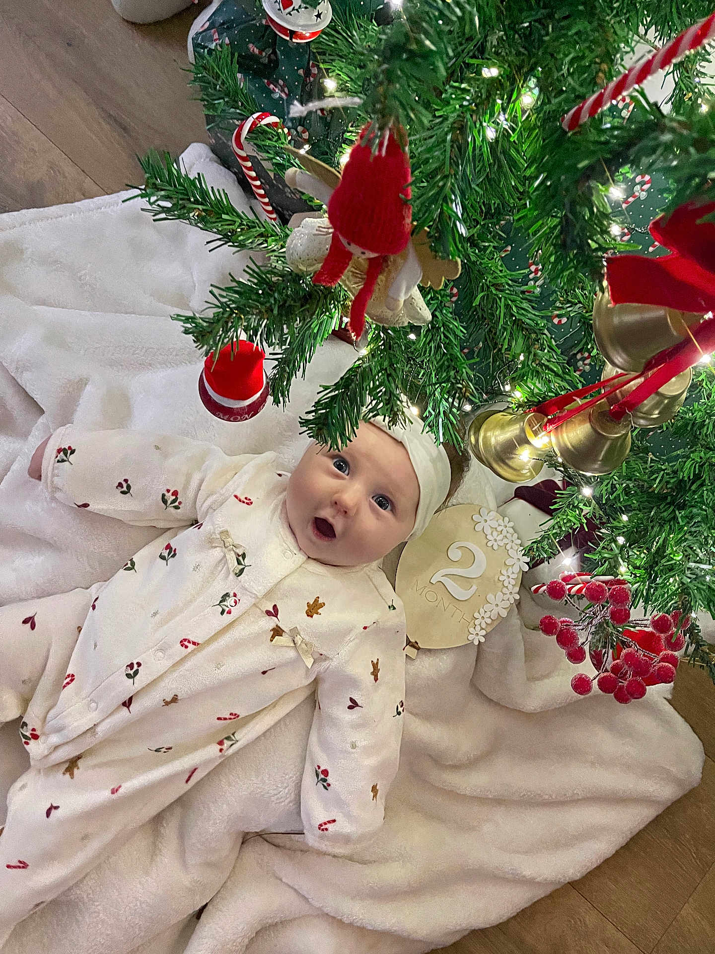 Dohya a rejoint le concours — aidez-le/la à gagner de superbes lots ! baby, infant, christmas_tree, ornaments, holiday_decor, blanket, white_clothing, headband, floor, wood_floor, greenery, bells, red_ornaments, lights, festive, cute, indoors, celebration, person, holiday