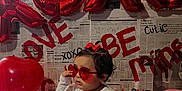 Gabby is registered to the contest to win money with this photo: toddler, child, balloons, red, heart_shaped_glasses, newspaper_background, valentines_day, love, xoxo, sitting, red_bow, white_shirt, red_pants, decorations, party, cute, indoors, celebration, smiling, person