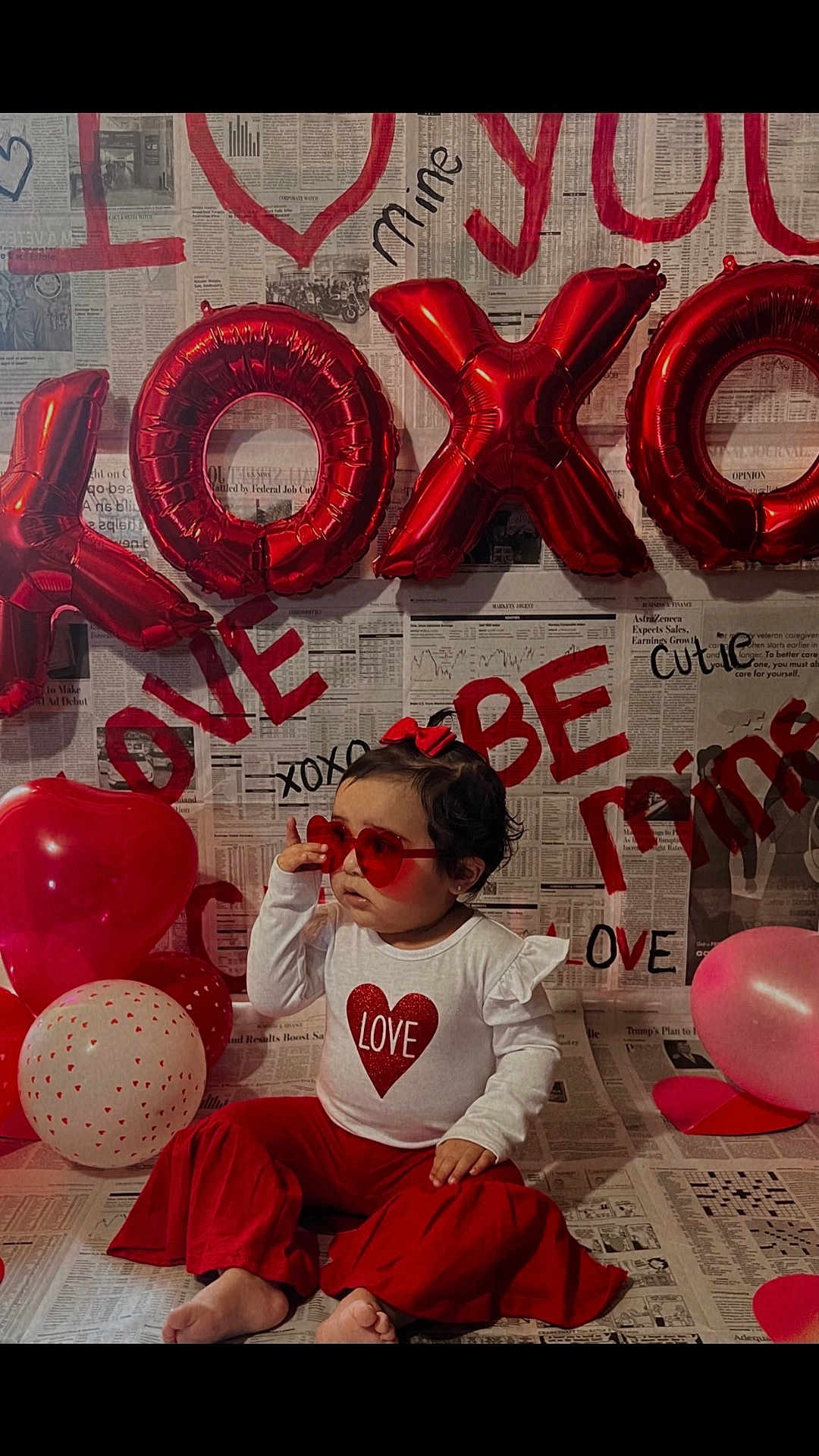 Gabby is registered to the contest to win money with this photo: toddler, child, balloons, red, heart_shaped_glasses, newspaper_background, valentines_day, love, xoxo, sitting, red_bow, white_shirt, red_pants, decorations, party, cute, indoors, celebration, smiling, person