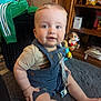 baby, child, infant, sitting, blue_overalls, beige_shirt, carpet, toy, stuffed_animal, shelf, indoor, cute, smiling, face, hand, foot, pacifier, wooden_furniture, curly_hair, bright_eyes