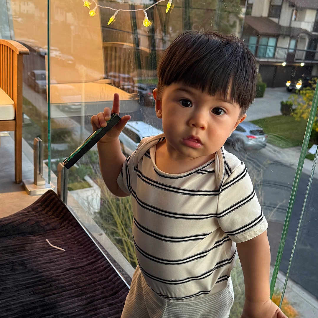 Cetin is registered to the contest to win money with this photo: balcony, car, child, curious, daylight, glass_railing, handrail, outdoor, pants, person, portrait, residential_area, socks, standing, star_lights, string_lights, striped_shirt, toddler, window, young_child