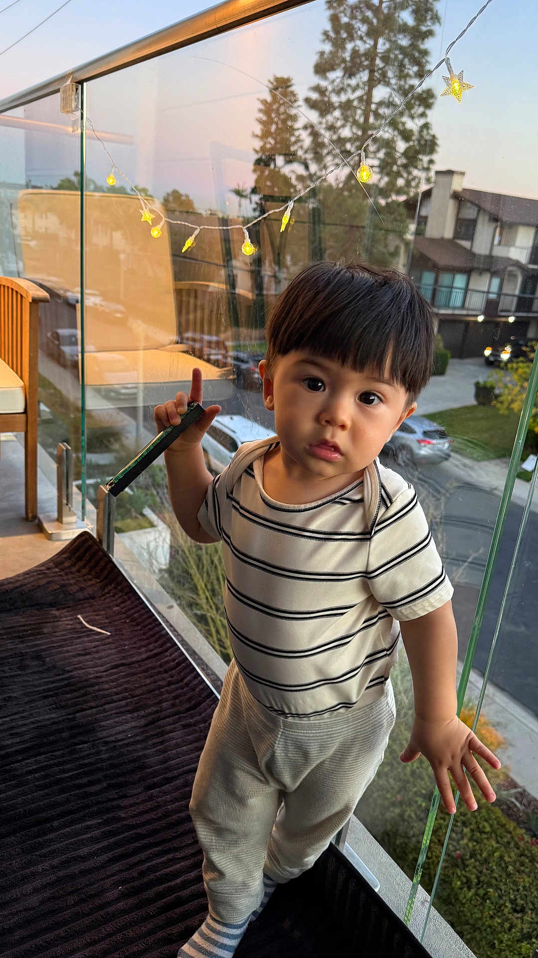Cetin is registered to the contest to win money with this photo: balcony, car, child, curious, daylight, glass_railing, handrail, outdoor, pants, person, portrait, residential_area, socks, standing, star_lights, string_lights, striped_shirt, toddler, window, young_child