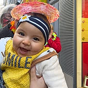 Loujayne participe au concours pour gagner de l'argent avec cette photo : adult, baby, child, clothing, colorful, face, hand, happy, hat, headband, headwear, outdoor, people, person, portrait, smiling, storefront, street, white_jacket, yellow_bib