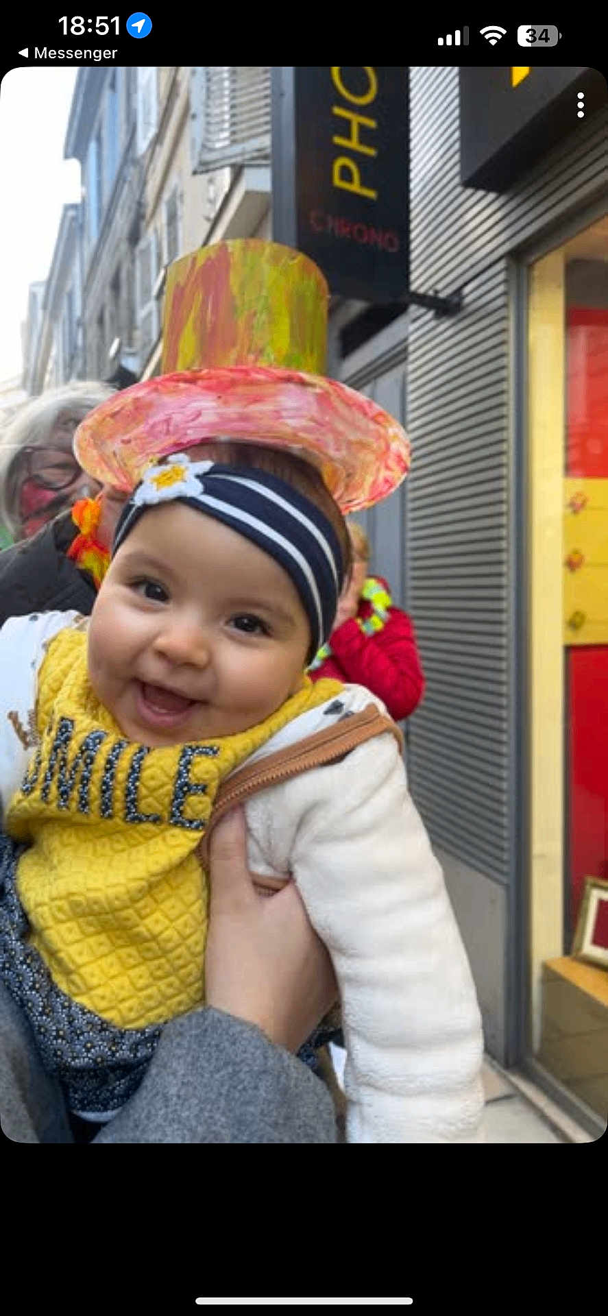 Loujayne participe au concours pour gagner de l'argent avec cette photo : adult, baby, child, clothing, colorful, face, hand, happy, hat, headband, headwear, outdoor, people, person, portrait, smiling, storefront, street, white_jacket, yellow_bib