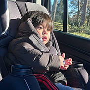 Rael is registered to the contest to win money with this photo: child, toddler, car_seat, winter_jacket, brown_hair, sunlight, window, trees, car_interior, cup_holder, seatbelt, looking_out, surprised, hands, bottle, sock, pants, travel, portrait, seat