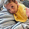 baby, infant, child, portrait, face, eyes, curious, indoors, blanket, bib, yellow_clothing, gray_bedding, lying_down, cozy, bedroom, close_up, brown_hair, sitting, home, photography