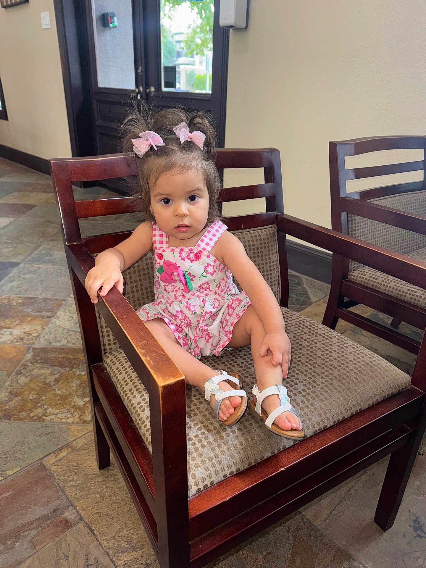 Sophia joined the competition — help win amazing prizes! toddler, child, girl, chair, wooden_furniture, pink_bows, floral_dress, sandals, indoor, floor_tiles, patterned_upholstery, curious_expression, sitting, hair_bows, casual_clothing, young_child, portrait, daylight, home_interior, cute