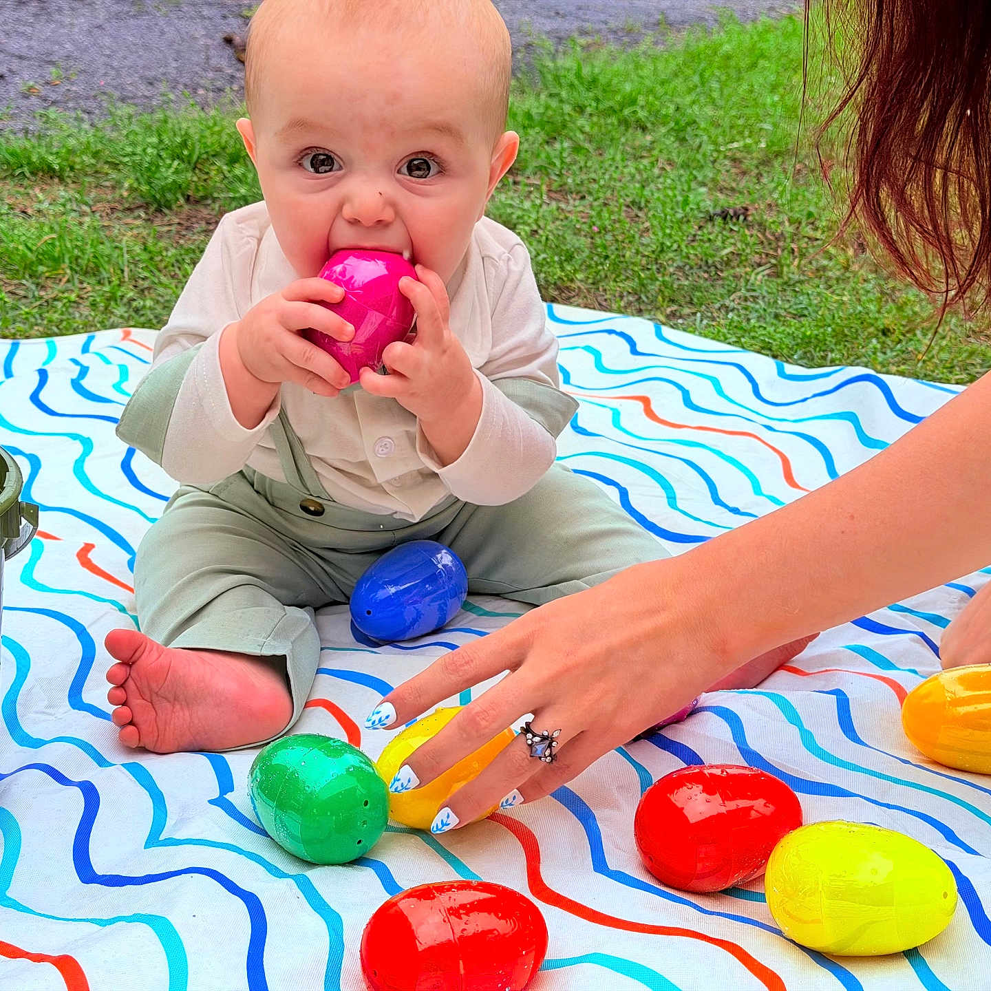 Gunner is registered to the contest to win money with this photo: baby, child, plastic_egg, colorful, outdoor, grass, blanket, hand, ring, nail_art, play, sitting, foot, green_bucket, toy, person, cute, bright_colors, chewing, fun
