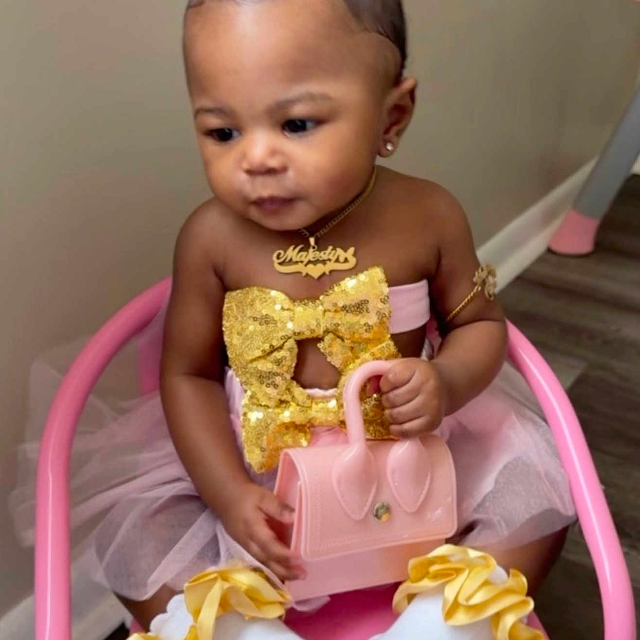 Majesty is registered to the contest to win money with this photo: baby, child, girl, pink_chair, pink_bow, gold_bow, tutu, pink_purse, white_socks, yellow_ruffles, jewelry, earrings, necklace, arm_band, sitting, indoor, flooring, cute, fashion, portrait