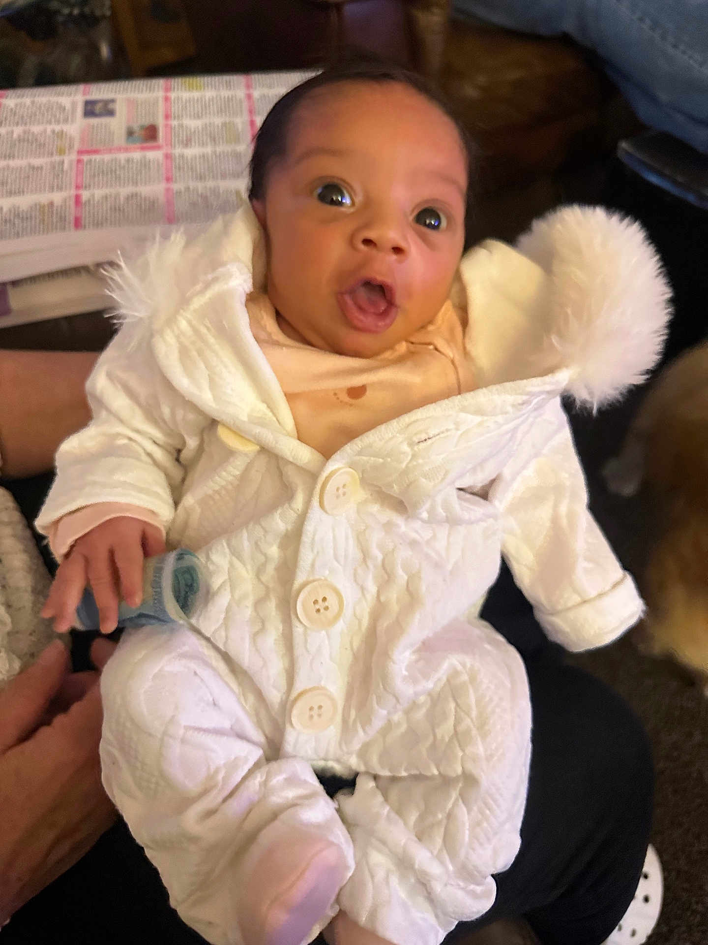 Aariellà is registered to the contest to win money with this photo: baby, child, infant, white_outfit, fur_trim, buttons, hand, person, indoors, newspaper, sitting, surprised_expression, warm_clothing, lap, cozy, pom_pom, face, eyes, mouth_open, holding