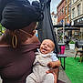 baby, woman, smiling, outdoor, cafe, urban, headwrap, glasses, happy, child, holding, clothing, street, table, chair, greenery, people, daylight, portrait, casual